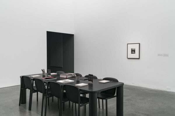 A black table with eight black chairs in the middle of a white room near a small artwork hung on the wall and a doorway covered in black fabric