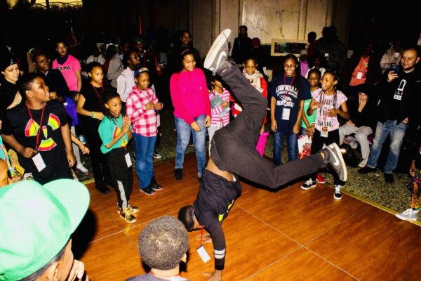 A breakdancer in a handstand freeze performs with a crowd of people of various ages around them.