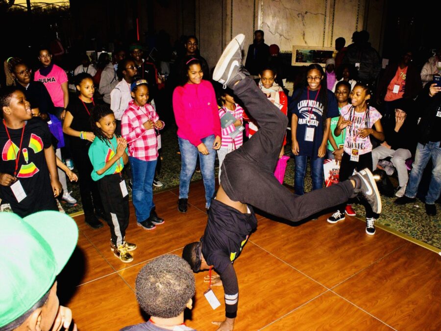 A breakdancer in a handstand freeze performs with a crowd of people of various ages around them.