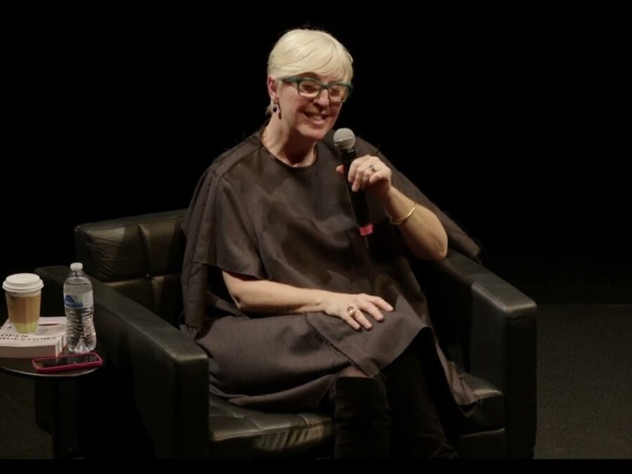 A woman with alabaster skin, dressed in all black with platinum blond hair, sits in a black chair on a black stage holding a microphone. A small table holds a coffee, water bottle, and book.