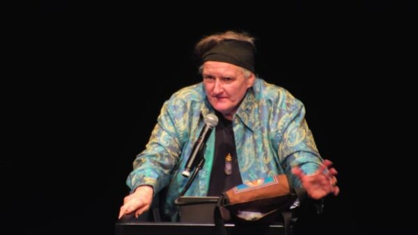 A person in a black headband and teal coat stands at a pedestal with a microphone in front of a black backdrop