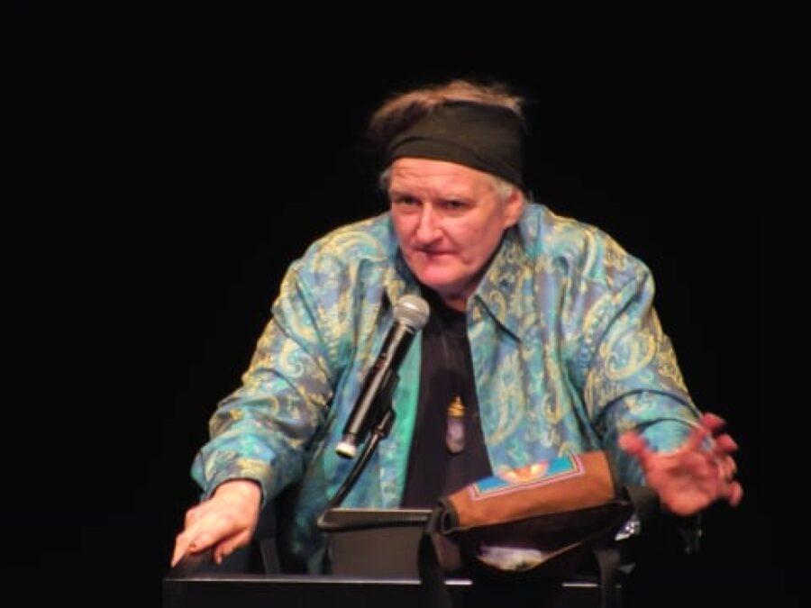 A person in a black headband and teal coat stands at a pedestal with a microphone in front of a black backdrop