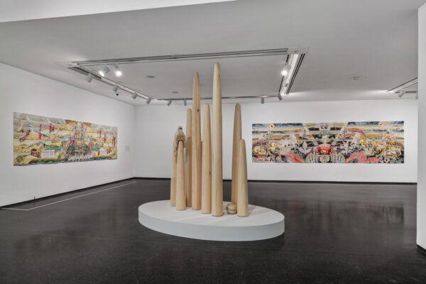 A series of various sized wooden spires on a white platform in the middle of a gallery space with two paintings hanging on two adjacent walls