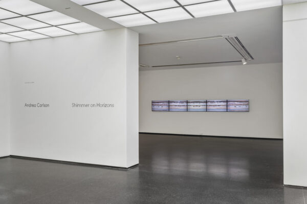 A wall with text reading Shimmer on Horizons next to a wide doorway with five screens placed side by side horizontally
