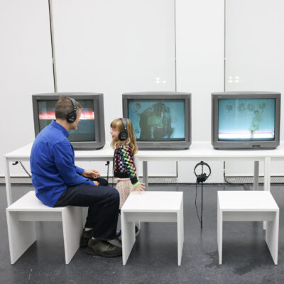 An adult male and a young child interact while sitting in front of four computers wearing headphones.