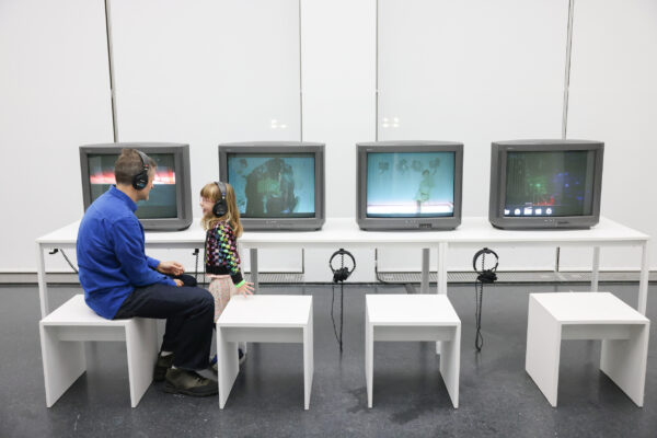An adult male and a young child interact while sitting in front of four computers wearing headphones.