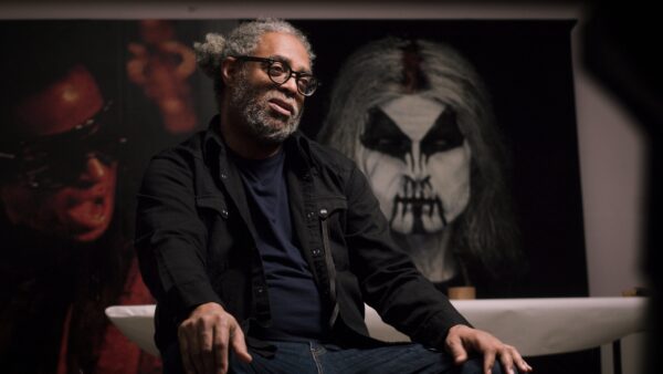 Arthur Jafa sits in a darkened room with artworks behind him