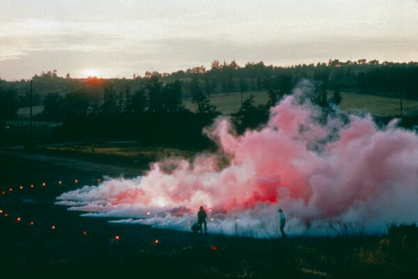 A pink cloud rises up from the ground in a darkened landscape with the sun barely appearing above a hill