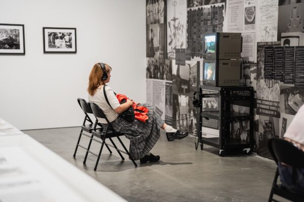 A person wearing headphones sits in a folding chair in front of a stack of four TVs