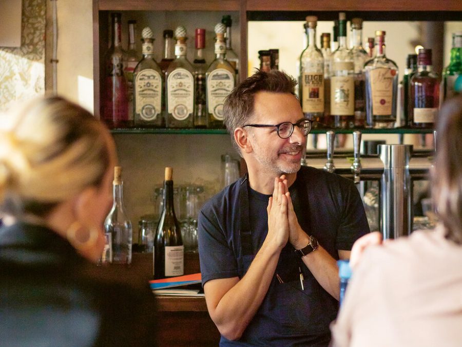 A man claps his hands together in front of a well-stocked bar as two people with their backs to the viewer look on