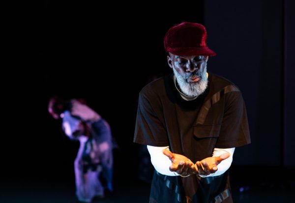 A man looks down at his outstretched, cupped hands as an out-of-focus figure bends forward in the background