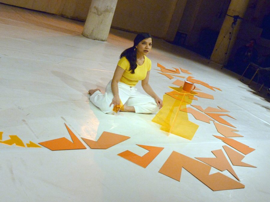 A woman sits on a sleek white floor among large geometric orange paper cutouts.