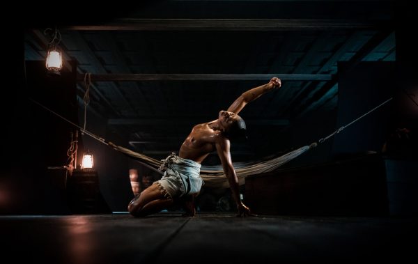 A topless person stretches out next to a hammock on a dimly lit stage.