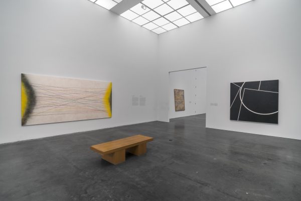 Gallery space with two paintings on a white wall and a bench in the middle of the room. A third painting is just visible through a doorway on the right wall