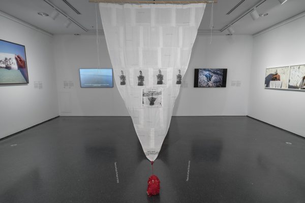 Gallery with several photographs, videos, and other artworks on the wall. In the center a sail-like object hangs from the ceiling.