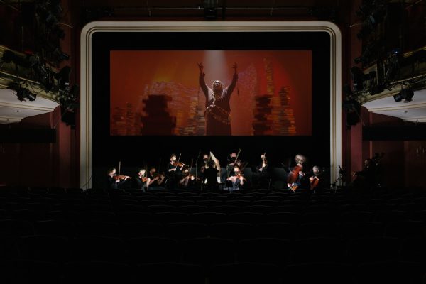 An orchestra plays in front of a screen displaying a film in a theater