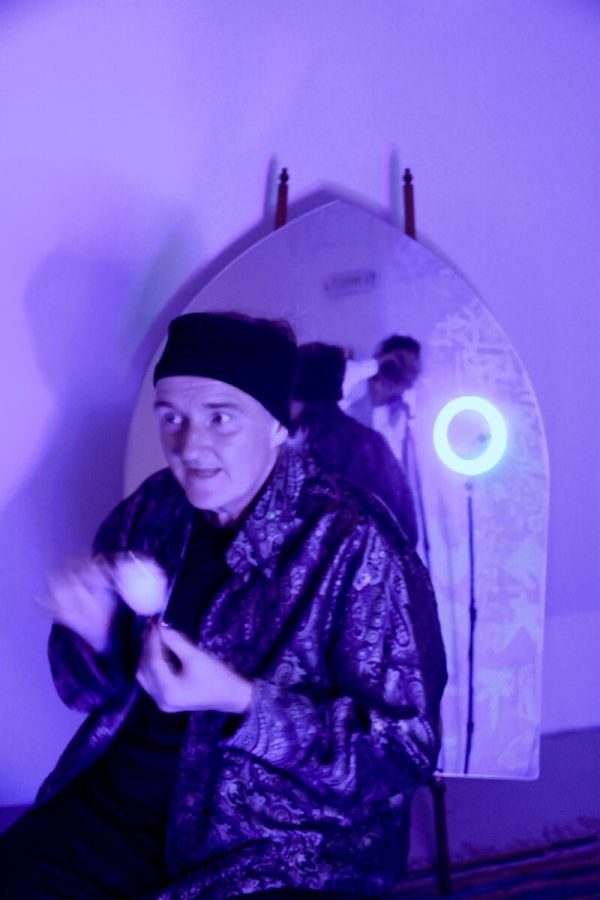 In a purple-lite room a person poses in front of a mirror revealing the photographer.