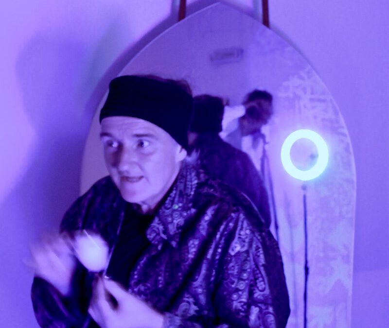 In a purple-lite room a person poses in front of a mirror revealing the photographer.