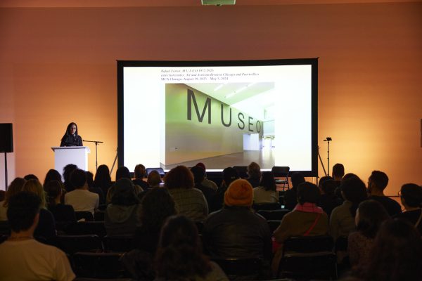 A crowd of people sit in front of a large presentation screen and a presenter at a podium in a dimly lit room.