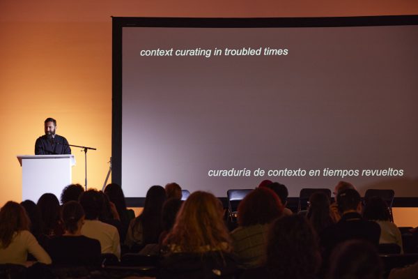A presenter at a podium beside a presentation screen with a crowd of people sitting facing them.