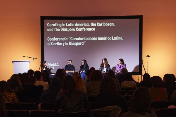 A panel of four people sit in front of a large presentation screen with an audience facing them.
