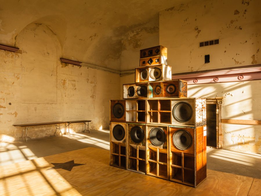 A stackful of various sized speakers in an otherwise empty room. Light streams from the windows, which cast shadows on the speakers, walls, and floor.