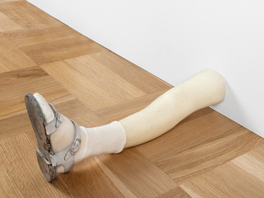 A prosthetic leg wearing a sandal and white sock rests on a wooden parquet floor against a white wall.