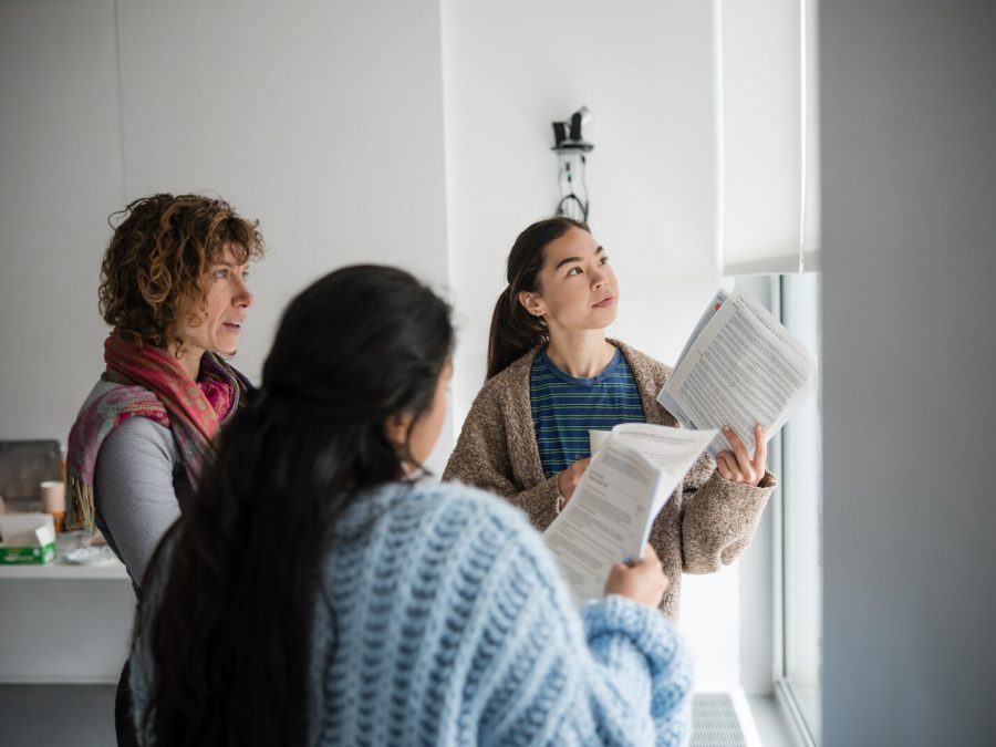 Three people standing in a semicircle look off into the distance. In their hands they hold a handful of paper.