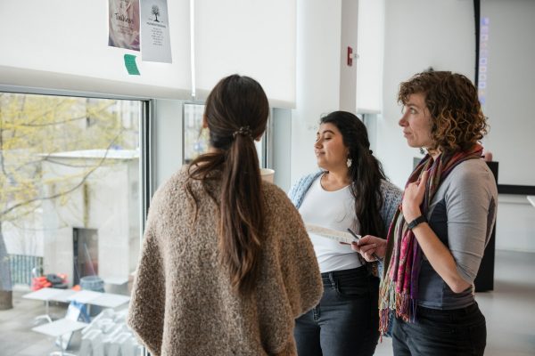Three people gather around pieces of paper taped to a window shade in a bright white studio space