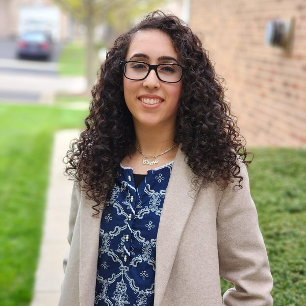 Three-quarter portrait of a female-presenting person with medium-light skin and curly dark hair that sits just past their shoulders and glasses posing outside