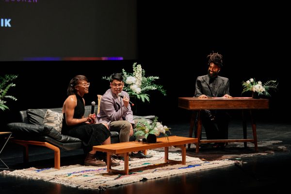 Two presenters sit in armchairs in front of a coffee table on a stage, while one person plays a keyboard.