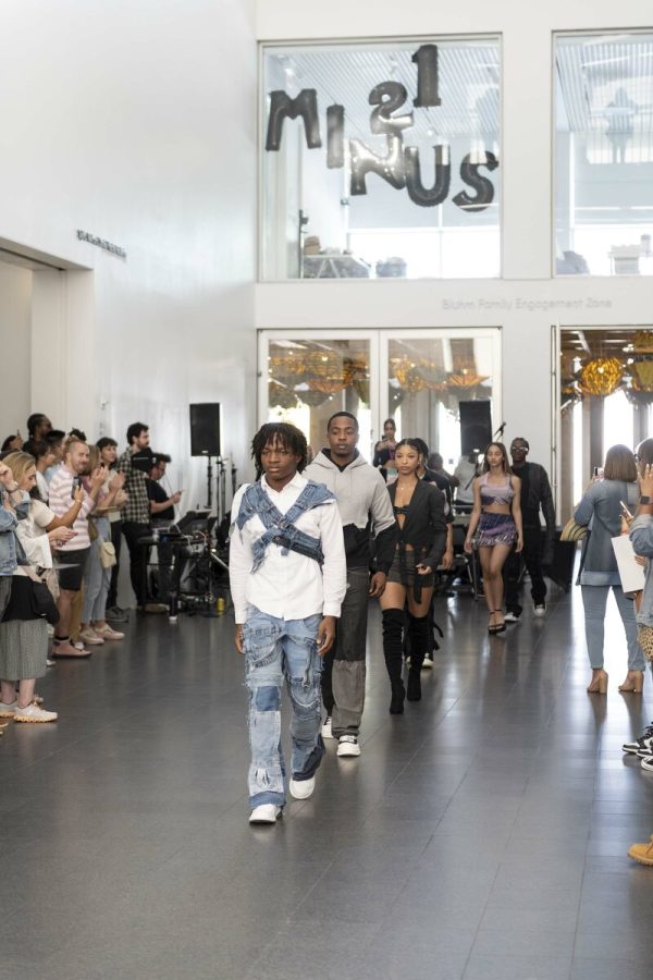A line of people walk in front of an audience in a large, airy white space.