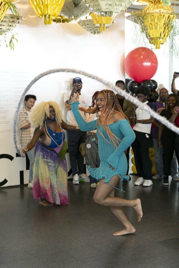 A female-presenting person in a teal dress jumps rope in a white room as a crowd watches behind them.