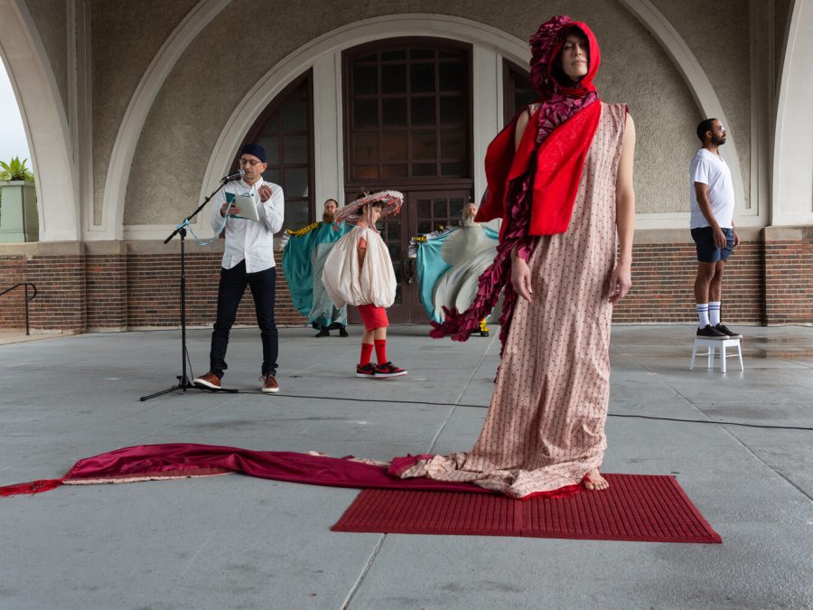 A person in a dress with a long train and red bonnet stands barefoot on a rug as a group of people in various costumes pose behind them