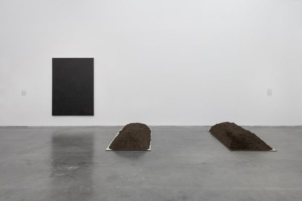 A white-walled gallery space with gray concrete flooring. Two artworks are visible: one appears to be a black canvas hung on the wall; the other consists of two mounds of earth, like two fresh graves.