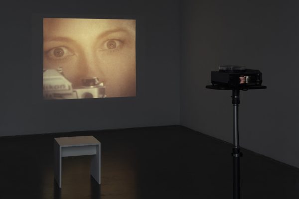 A dark room featuring a white stool and a projector is illuminated by a projection of a close-up of a woman's face with the top of a Nikon camera placed close to her eye.