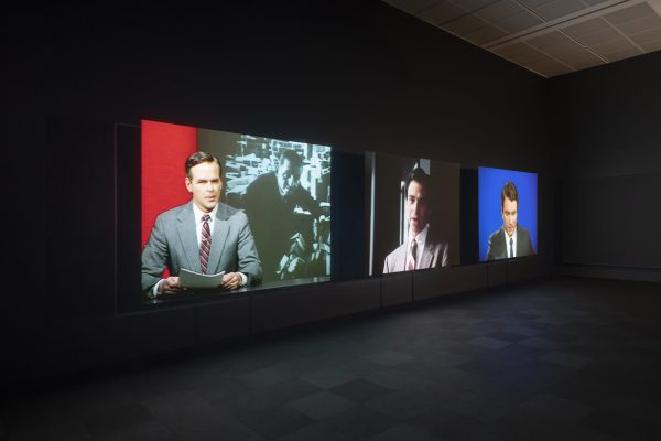 A dark room slightly illuminated by three projections of three newscasters.