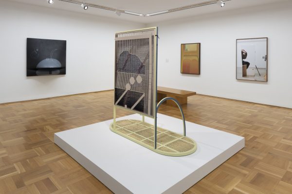 Four artworks on display in a white-walled gallery space. Three hang on the wall; one is installed on a pedestal.