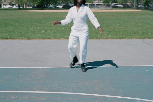 A person skips in a white jumpsuit on a court in a park setting, their head is not visible from the lips up