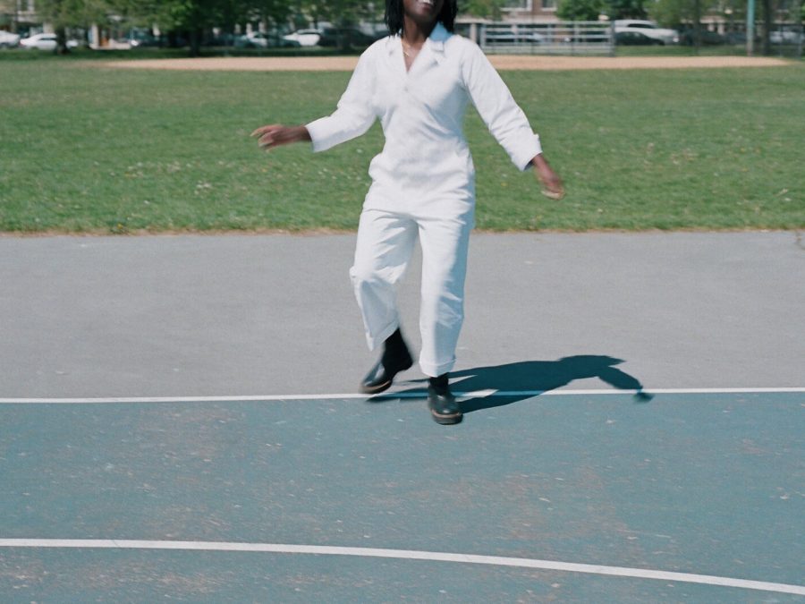 A person skips in a white jumpsuit on a court in a park setting, their head is not visible from the lips up
