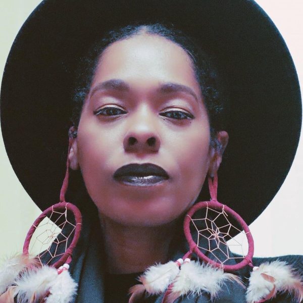 Close up stylized portrait of a female-presenting person with medium skin wearing a dark wide-brimmed hat and red dream catcher earrings