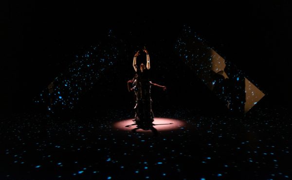 A person is spotlit on a black stage surrounded by three projections of little blue lights. Another person's arms are just visible behind the front actor.