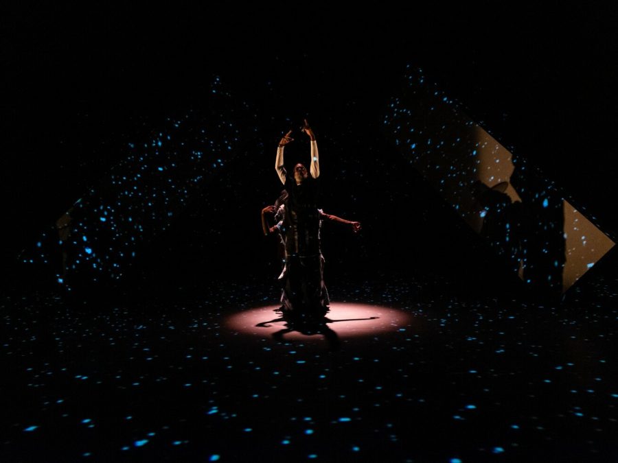 A person is spotlit on a black stage surrounded by three projections of little blue lights. Another person's arms are just visible behind the front actor.