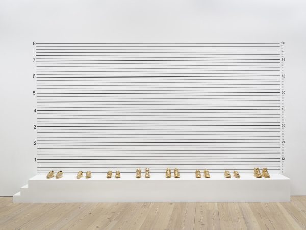 Photo of 8 pairs of gold-plated basketball shoes evenly spaced out on a white platform in front of a white wall with black numbers and lines.
