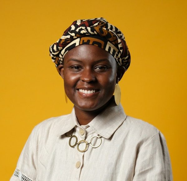 A smiling female-presenting person with dark skin wearing a headscarf poses in front of a mustard yellow backdrop