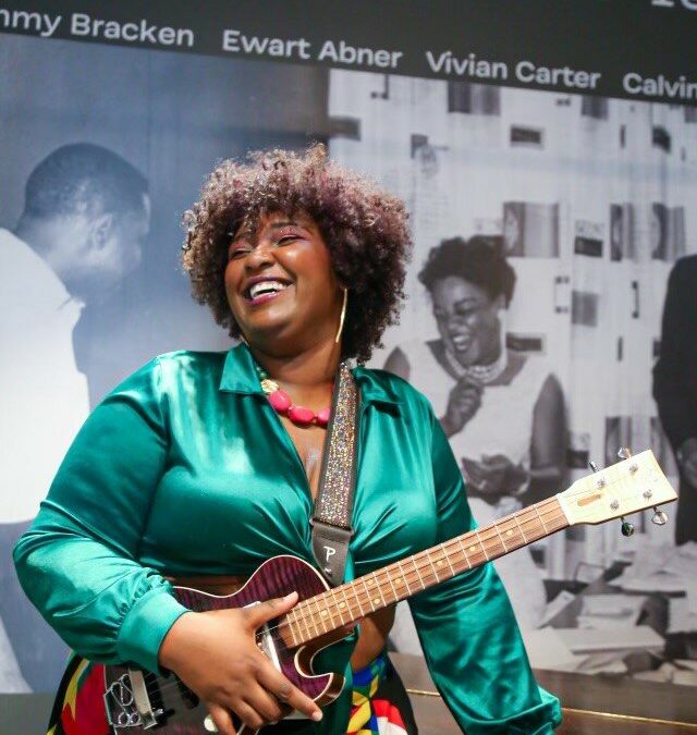 Photo of a brightly dressed Black woman standing in front of black-and-white posters labeled Vee-Jay Records. Her left hand rests on a wooden table while her right hand holds onto a guitar. She smiles broadly towards something off camera.