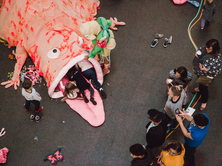 A group gathers near a large pink monster that has a few pairs of legs hanging out of its mouth