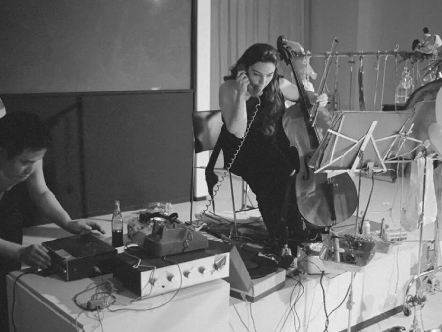 Archival photo of two people interacting with various sound-making objects, including balloons, a cello, and a record player, while one person holds a telephone receiver to their ear.