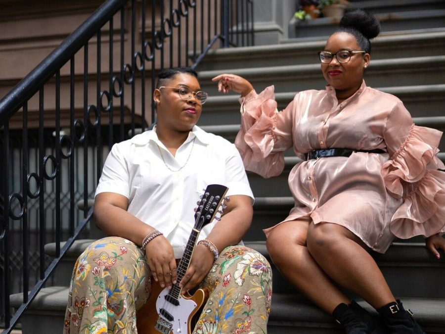 Two Black femmes sitting on the stairs of a brownstone.