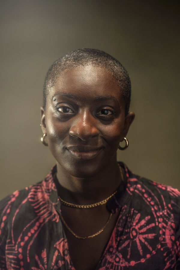 Portrait of a female-presenting person with dark skin and very short dark hair wearing a black and hot pink patterned shirt against a grey backdrop